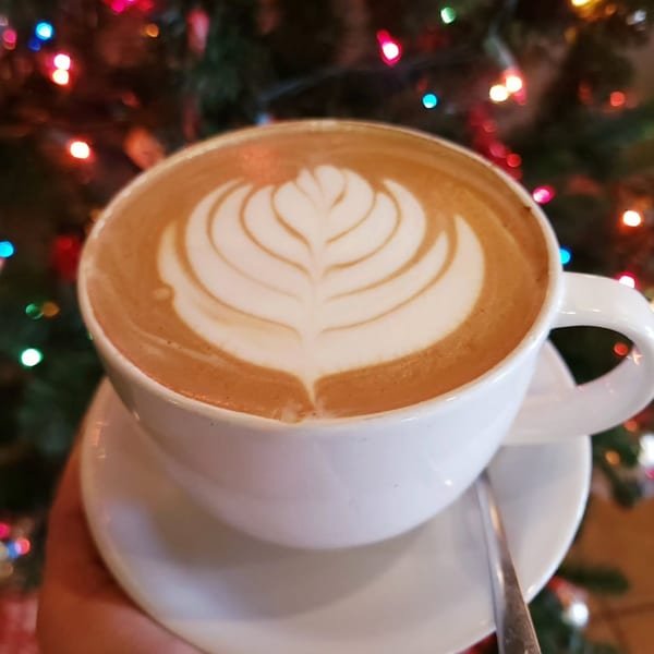 A latte with intricate foam art in a white cup, held against a blurred background of festive lights.