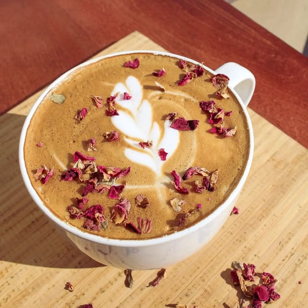 A latte with a leaf design in the foam, topped with dried rose petals, on a wooden surface.